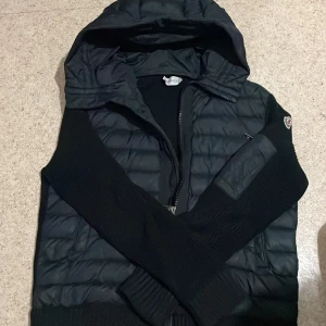 Svart pufferjacka från Moncler - Svart pufferjacka från Moncler med huva och stickade ärmar. Jackan har quiltad framsida, dragkedja och Moncler-logga på ärmen. Materialmix av polyester och stickad textil ger en schysst kontrast. Perfekt för dig som vill ha en snygg och varm jacka med street vibe.