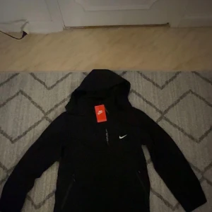 Svart Nike vindjacka med huva - Nike vindjacka i svart med huva och vit logga på bröstet. Jackan har dragkedja framtill och trycket 'EXPLORE' längs dragkedjan. Snygg och sportig design, perfekt för dig som gillar streetwear och vill ha något stilrent från Nike.