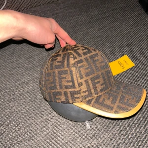 Fendi keps med FF-logga, brun - Snygg keps från Fendi med ikoniskt FF-mönster i mörkbrunt och svart. Kepsen har en böjd skärm med beige kant och justerbar rem baktill i skinn med Fendi-logga. Materialet är bomull och skinn, perfekt för dig som vill sticka ut med designerstil.
