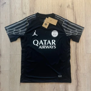 Svart PSG x Jordan fotbollströja S - Snygg svart PSG fotbollströja från Jordan med vita detaljer och tryck. Tröjan har korta ärmar med unikt mönster, rund hals och är gjord i lätt Dri-Fit material som andas. Ikoniska PSG-loggan och Jumpman på bröstet samt Qatar Airways tryck.