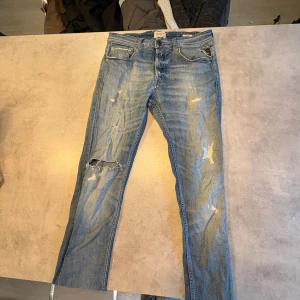 Replay blå jeans med slitningar - Snygga blå jeans från Replay med coola slitningar och hål på benen för en avslappnad streetstil. Klassisk femficksmodell, ljus tvätt och raka ben. Jeansen har Replay-logga på fickan och brunt läderpatch bak i midjan. Perfekta för dig som gillar en edgy look.