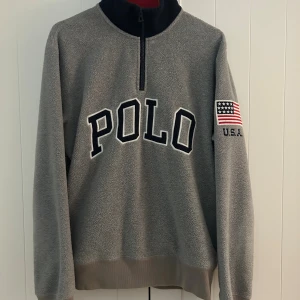Grå fleece-tröja Polo Ralph Lauren - Grå fleece-tröja från Polo Ralph Lauren med svart krage och röd insida. Stor POLO-text på bröstet och amerikansk flagga med U.S.A. på vänster ärm. Half zip framtill och ribbad nederkant. Perfekt för kyliga dagar och riktigt skön att ha på sig. Liten skada på rygg, annars som ny!