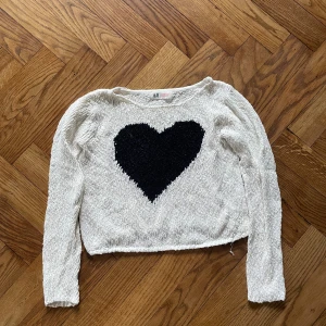 Vit stickad tröja med svart hjärta H&M - Superfin vit stickad tröja från H&M med ett stort svart hjärta på bröstet. Tröjan har lång ärm och är gjord i 100% bomull, vilket gör den både mjuk och skön. Perfekt för dig som gillar gulliga detaljer och vill ha en mysig look.
