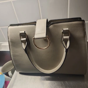 Beige handväska med gulddetaljer - Snygg beige handväska med stilrena guldfärgade ringdetaljer och praktiska handtag. Väskan har flera fack och dragkedja upptill för smidig organisering. Tillverkad i skinnimitation med svart kant och elegant, strukturerad form. Perfekt för dig som gillar klassisk och clean stil.