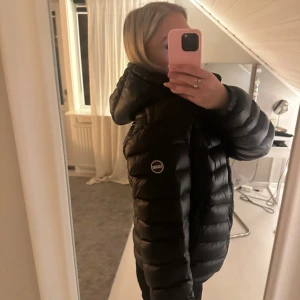 Svart Colmar dunjacka med huva - Svart dunjacka från Colmar med huva och diskret logga på ärmen. Jackan har en puffad look, dragkedja framtill och två sidofickor. Passformen är normal och jackan är perfekt för kalla dagar. Stilren och enkel design som funkar till många outfits. Pris kan justeras! Har en lapp på ena ärmen (som man ser på en av bilderna) men den är väldigt väldigt diskret så inget som sticker ut!