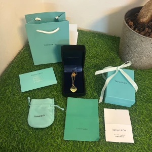 Guldigt hjärthalsband Tiffany & Co - Exklusivt halsband från Tiffany & Co i 18K guld med ikonisk hjärtberlock och ingraverad text 'Please return to Tiffany & Co New York 750'. Kedjan är tunn och elegant, och halsbandet levereras med originalask, påse och certifikat. Perfekt statement-piece för dig som älskar lyxiga smycken.