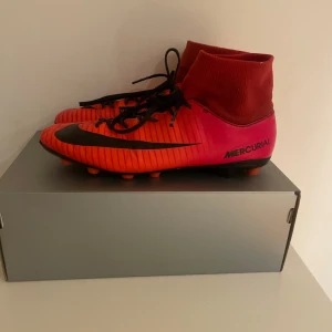 Nike Mercurial fotbollsskor röd/svart - Säljer ett par Nike Mercurial fotbollsskor i rött och svart med högt skaft och snörning. Skorna har en snygg design med stora Nike-loggor på sidan och är perfekta för dig som vill sticka ut på planen. Sulan har dobbar för bra grepp på gräs.