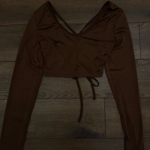Brun croppad topp med snörning bak - Säljer en brun långärmad croppad topp med djup v-ringning och snygga korsade band i ryggen. Toppen har en cool snörningsdetalj baktill som ger en rynkad effekt och extra edge. Perfekt för dig som gillar trendiga och unika plagg.