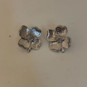 Silvriga blomformade örhängen - Ett par snygga örhängen i form av blommor med detaljerade kronblad. De är i en blank silvrig färg och har en tydlig blomdesign som sticker ut. Perfekta för dig som vill ha något unikt och stilrent i öronen. Köpte två par av samma öronhänge så dessa är aldrig använda 