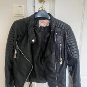 Svart skinnjacka från Voyelles, stl 36 - Snygg svart skinnjacka från Voyelles med coola detaljer som quiltade axlar, dragkedjor och bälten i nederkant. Jackan har klassisk bikerstil med asymmetrisk dragkedja och markerad krage. Perfekt för dig som vill ha en edgy look.    Nästan aldrig använd. Nypris: 699kr