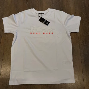 Vit t-shirt från Hugo Boss - Vit t-shirt från Hugo Boss med broderad logga i vitt och röd text på bröstet. Klassisk rund halsringning och korta ärmar. Tillverkad i mjuk bomull för en skön känsla. Perfekt för en clean och stilren look.