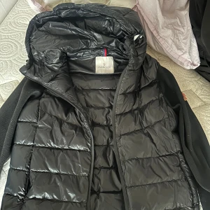 Svart Moncler cardigan - Snygg svart pufferjacka från Moncler med glansig finish och huva. Jackan har quiltad framsida och rygg, samt stickade svarta ärmar med Moncler-logga på ena ärmen. Perfekt för kyliga dagar och riktigt stilren look.