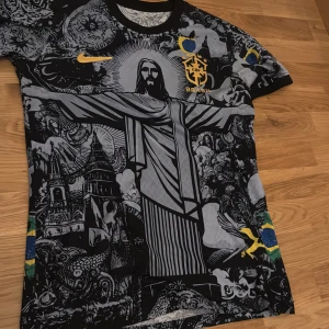 Fotboll T-shirt  - Unik Brasilien fotbollströja från Nike med stort tryck av Kristusstatyn i Rio och detaljerade svartvita illustrationer. Gula Nike-loggan och Brasilien-emblem på bröstet, samt brasilianska flaggan på 