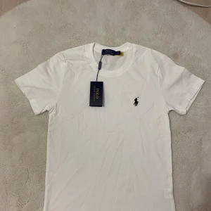 Vit t-shirt från Polo Ralph Lauren - Vit klassisk t-shirt från Polo Ralph Lauren med rund hals och korta ärmar. Ikonisk broderad logga i svart på bröstet. Tillverkad i mjuk bomull för en skön känsla. Perfekt basic-plagg med snygg passform och stilren look.