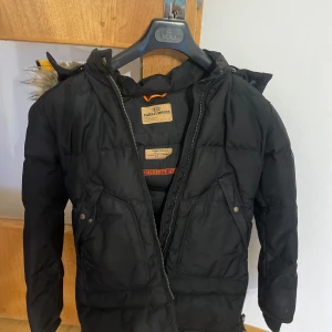 Svart dunjacka Parajumpers med päls - Svart dunjacka från Parajumpers med huva fodrad i mjuk fuskpäls och snygg pälskant. Jackan har dragkedja, stora fickor framtill och coola detaljer som orange hängare i nacken. Perfekt för kalla dagar när du vill hålla stilen.