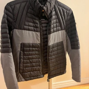 Svart och grå dunjacka från Hugo Boss - Snygg dunjacka från Hugo Boss i svart och grått med quiltad design. Jackan har dragkedja framtill och två stora fickor. Märkeslogga på ärmen och lätt vaddering. Jackan är i bra skick, bara hängaren som är trasig. Pris kan diskuteras 🌸