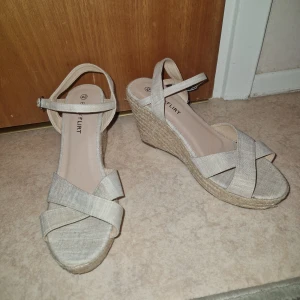 Kilklackar  - Snygga beiga sandaler från Bodyflirt med kilklack i rep och breda korslagda remmar. Skorna har öppen tå, justerbart spänne vid ankeln och ovandel i textil. Perfekta för sommarens alla tillfällen när du vill ha lite höjd men ändå bekvämt.