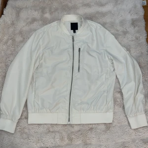Vit bomberjacka från H&M, storlek L - Snygg vit bomberjacka från H&M i storlek L. Jackan har klassisk bomberkrage, dragkedja framtill och en bröstficka med dragkedja. Tillverkad i blankt syntetmaterial med ribbade muddar och foder i satin-look. Perfekt för dig som gillar stilren och clean look.