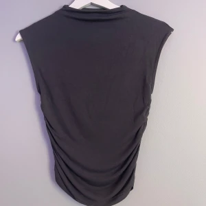 Mörkgrå ärmlös topp från Gina Tricot - Säljer en mörkgrå ärmlös topp från Gina Tricot i mjukt och stretchigt material. Toppen har en enkel och stilren design med rak halsringning och lätt rynkade sidor som ger en snygg passform. Perfekt att matcha med jeans eller kjol.