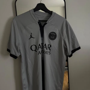 PSG grå fotbollströja Jordan - Snygg grå Paris Saint-Germain fotbollströja från Jordan med svart logga och sponsortryck. Tröjan har en svart vertikal rand i mitten, korta ärmar och svarta detaljer vid ärmslut och krage. Perfekt för dig som älskar fotboll och streetwear.