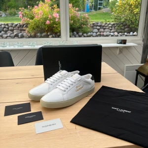 Vita Saint Laurent sneakers - Snygga vita sneakers från Saint Laurent med guldfärgad logotyp på sidan och svart detalj vid hälen. Skorna är tillverkade i lyxigt skinn och har klassisk snörning samt en platt, beige sula. Perfekt för dig som gillar stilrena och exklusiva sneakers. Box, dustbag och kort från Saint Laurent ingår. Säljer då de var för små för mig! Bara att skriva vid funderingar😁