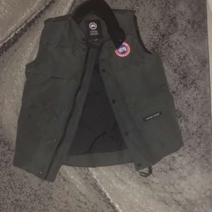 Grå  dunväst från Canada Goose XS - Säljer en svart dunväst från Canada Goose i storlek XS. Västen har klassisk quiltad design, hög krage och tryckknappar framtill. Ikonisk Canada Goose-logga på bröstet och tydlig märkning på fickan. Perfekt för kalla dagar och snygg att styla över hoodie.