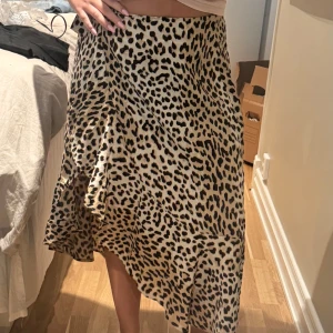 Leopardmönstrad långkjol i satin - Snygg långkjol med leopardmönster i beige, svart och bruna toner. Kjolen har en lätt glansig satinliknande yta och faller snyggt med en lite asymmetrisk nederkant. De är en knytning i midjan. Perfekt för dig som vill sticka ut med ett djurmönster och gillar flowiga kjolar.