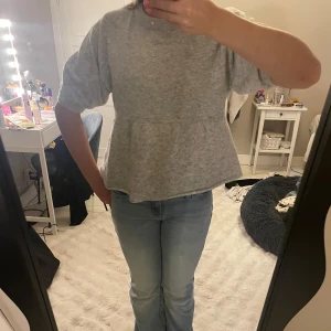 Grå stickad topp från Nelly - Supermysig grå stickad topp från Nelly med korta puffärmar och rund hals. Modellen har en lätt utsvängd peplum nedtill som ger en söt siluett. Perfekt till jeans eller kjol för en chill look.