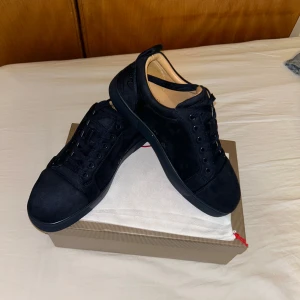 Svarta sneakers från Christian Louboutin - Snygga svarta sneakers från Christian Louboutin i mocka med klassisk röd sula. Skorna har rund tå, svarta snören och diskreta detaljer. Insidan är beige och de har en stilren, minimalistisk look som passar till det mesta.