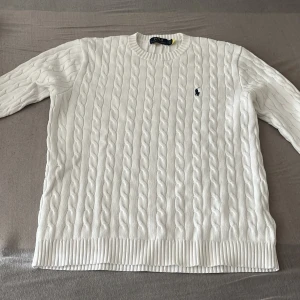Vit kabelstickad tröja från Polo Ralph Lauren - Säljer en vit kabelstickad tröja från Polo Ralph Lauren med klassisk rund hals och broderad marinblå logga på bröstet. Tröjan har långa ärmar och ribbade muddar vid hals, ärmslut och nederkant. Perfekt för dig som gillar stilren och tidlös design.