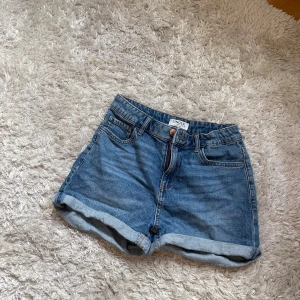 Blå jeansshorts från Lindex - Snygga blå jeansshorts från Lindex med klassisk femficksdesign och uppvikta benslut. Shortsen har normal passform, bälteshällor och stängs med knapp och dragkedja. Perfekta för varma dagar och har en tidlös look som funkar till allt.