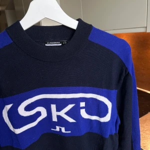 Blå och svart stickad tröja J.Lindeberg - Säljer en snygg stickad tröja från J.Lindeberg i svart och blått med vit SKI-logga framtill. Tröjan har rund halsringning, långa ärmar och ribbade muddar. Perfekt för after ski eller i fjällvärlden! Nypris 1900kr och den är knappt använd. Säljer för 800kr 