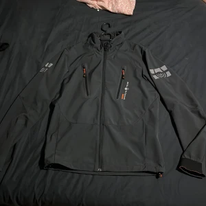 Svart vindjacka från Sail Racing - Svart vindjacka från Sail Racing med hög krage, två bröstfickor med dragkedja och snygga loggor på ärmarna och ryggen. Jackan har orange detaljer vid dragkedjorna och är tillverkad i ett slitstarkt syntetmaterial. Perfekt för blåsiga dagar och sportig stil.