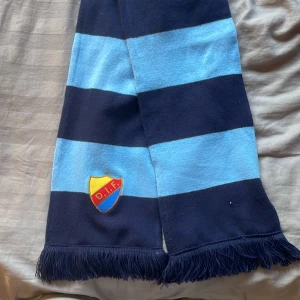 Randig Djurgården halsduk blå/marin - Snygg halsduk från Djurgårdens IF med breda marinblå och ljusblå ränder, broderad DIF-logga och fransar i ändarna. Perfekt accessoar för att visa ditt stöd på matchen eller i vardagen. Köpt på stadium💞