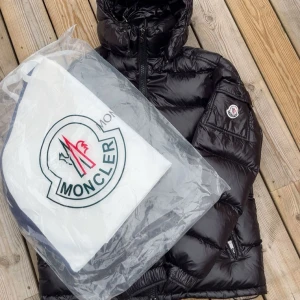 Moncler Maya - Säljer en svart pufferjacka från Moncler med glansig finish, huva och dragkedja framtill. Jackan har Moncler-logga på ärmen och praktisk ficka med lock. Insidan har ett unikt serietidningsmärke. Perfekt för kalla dagar och riktigt snygg streetstil. Size 2=M. Dustbag och tags följer med.