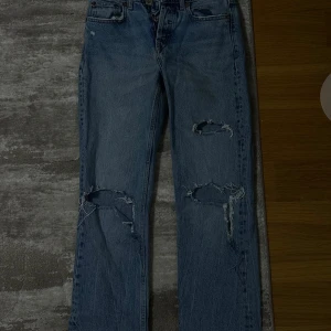 Blå slitna raka jeans - Säljer ett par blå jeans med raka ben och slitna detaljer på båda benen. Klassisk femficksmodell med knappgylf och coolt vintage-vibe. Perfekta för en avslappnad streetstyle-look.