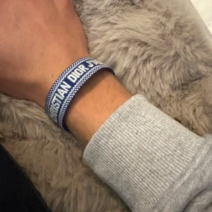 Blått Christian Dior J'Adior armband - Säljer ett snyggt blått armband från Christian Dior med texten 'J'Adior' broderad i vitt. Armbandet är gjort i textil och har tofsar i ändarna för en cool och avslappnad look. Perfekt accessoar för att lyfta din outfit.