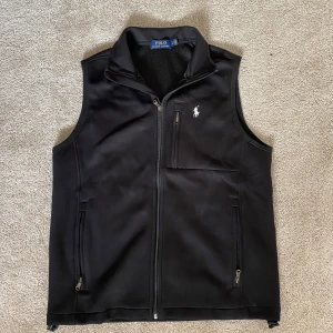 Svart väst från Polo Ralph Lauren - Svart väst från Polo Ralph Lauren med dragkedja framtill och flera fickor med dragkedja. Klassisk design med hög krage och meshfoder på insidan. Diskret logga broderad på bröstet. Perfekt för lager-på-lager och stilren look.