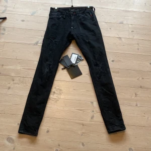 Svarta Philipp Plein slim jeans med döskalle - Svarta slim fit jeans från Philipp Plein med slitna detaljer och broderad döskalle på bakfickan. Jeansen har klassisk femficksdesign, små färgdetaljer vid fickan och metallknapp med logga. Snygg och edgy look med exklusiv känsla. Självklart äkta. Tillbehören följer med vid köp. Hör av er vid minsta lilla