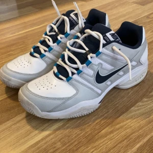 Nike vita och marinblå sneakers - Nike sneakers i vitt och marinblått med grå detaljer och blå accenter. Klassisk design med snörning, rund tå och platt sula. Ovandel i syntet och skinn, vadderad insida för extra komfort. Perfekta för dig som gillar sportig stil och vill ha ett par snygga och bekväma skor.