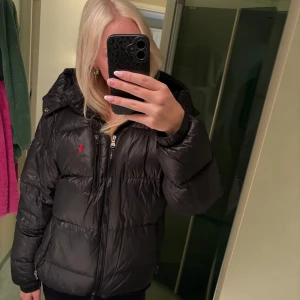 Svart pufferjacka från Polo Ralph Lauren - Svart pufferjacka från Polo Ralph Lauren med röd broderad logga på bröstet. Jackan har huva, dragkedja framtill och två sidofickor med dragkedja. Fylld för extra värme och har en klassisk, lite boxig passform.