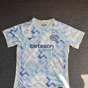 Inter Milan fotbollströja Nike blå/vit - Inter Milan fotbollströja från Nike med geometriskt mönster i ljusblått, vitt och turkost. Tröjan har korta ärmar, rund hals och klubbmärke samt sponsortryck på bröstet. Tillverkad i lätt och ventilerande funktionsmaterial.