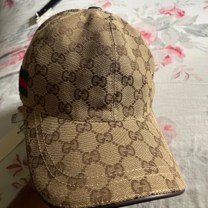 G keps med GG-mönster och läderdetalj - Snygg beige keps från  med klassiskt GG-monogram över hela ytan. Kepsen har bruna och gröna detaljer på sidan samt justerbar rem i mörkbrunt läder baktill. Tillverkad i bomull och läder, perfekt för dig som vill sticka ut med lyxig streetstyle.