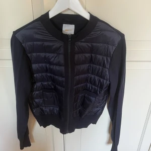 Svart quiltad kofta från Gina Tricot - Snygg svart kofta från Gina Tricot med quiltad framsida och dragkedja. Långärmad modell med ribbade muddar och två fickor framtill. Perfekt för lager-på-lager under höst och vinter.