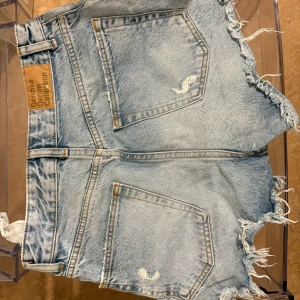 Ljusa jeansshorts från Bershka - Säljer ett par ljusblå jeansshorts från Bershka Denim Collection. De har hög midja, slitna detaljer och fransiga benslut för en cool vibe. Klassisk femficksmodell med dragkedja och knapp framtill. Perfekta för varma dagar.