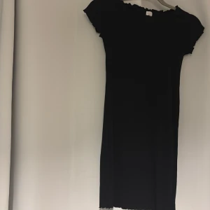 Svart ribbad kortklänning Lindex - En svart ribbad kortklänning från Lindex med korta ärmar och vågig kant vid hals och ärmslut. Klänningen har en enkel, tight passform och är tillverkad i mjukt stretchigt material. Perfekt för dig som gillar en stilren och basic look.