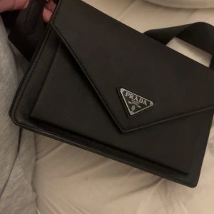 Svart axelväskai skinn - Säljer en svart axelväska  i stilren design med triangelformad logga framtill. Väskan har justerbar axelrem i textil, flera fack inuti och stängs med lock. Tillverkad i strukturerat skinn och har silvriga metalldetaljer.