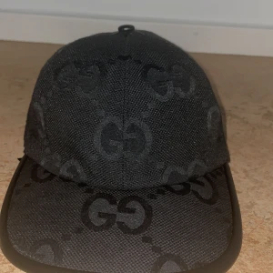 Svart keps från Gucci med GG-mönster - Snygg svart keps från Gucci med klassiskt GG-monogram över hela kepsen. Kepsen har böjd skärm, justerbart spänne bak i skinn och är tillverkad i ett slitstarkt tyg. Perfekt för dig som vill ha en stilren och lyxig accessoar.