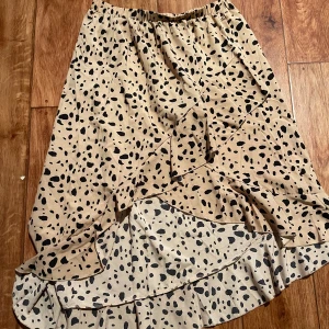 Beige kjol med svart leopardmönster - Säljer en beige långkjol med svart leopardmönster och volangdetaljer. Kjolen har resår i midjan och är tillverkad i ett lätt och mjukt tyg som ger ett snyggt fall. Perfekt för dig som gillar trendiga mönster och vill sticka ut.