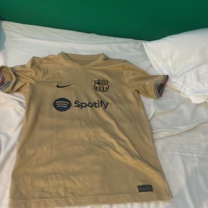 FC Barcelona guld fotbollströja Nike L - FC Barcelona fotbollströja i guld från Nike med Dri-FIT-material. Tröjan har korta ärmar med färgade detaljer vid muddarna, klubbmärke och Nike-logga på bröstet samt Spotify som sponsortryck. Perfekt för dig som älskar Barca och vill sticka ut på planen.